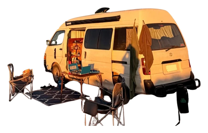 Hank at sunset with the side door open, two camp chairs, a table, gas stove and outdoor mat set up